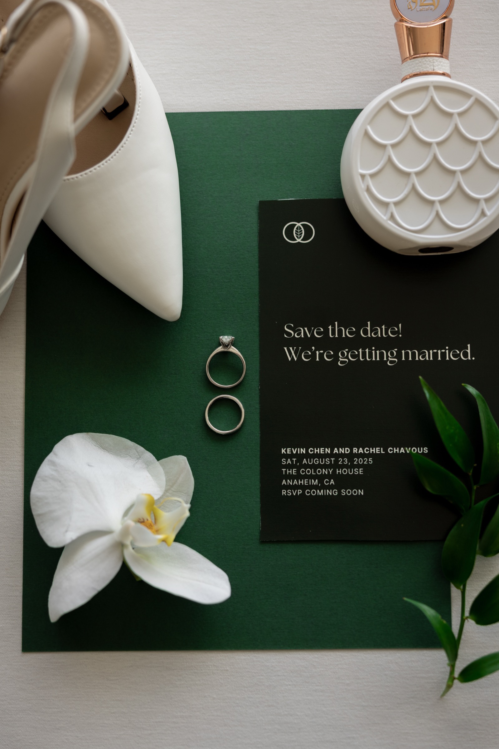 Wedding flat lay with save the date invitation, rings, white bridal shoes, orchid, and perfume for Kevin Chen and Rachel Chavous wedding at The Colony House