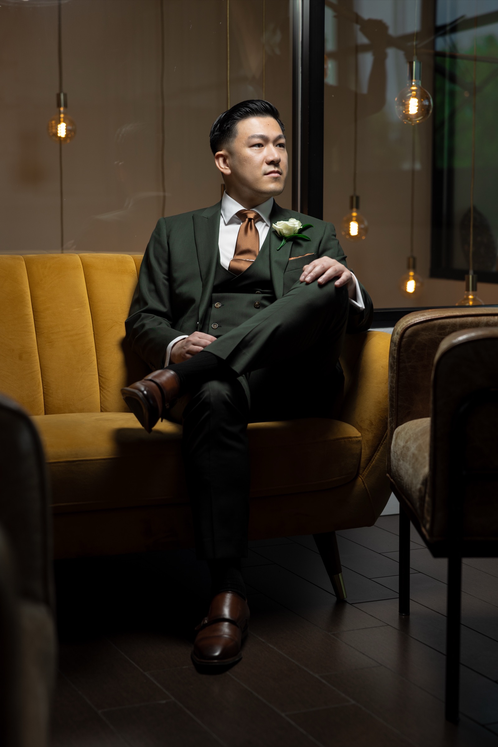 Kevin seated on yellow velvet couch with pendant lights, groom portrait at The Colony House Anaheim