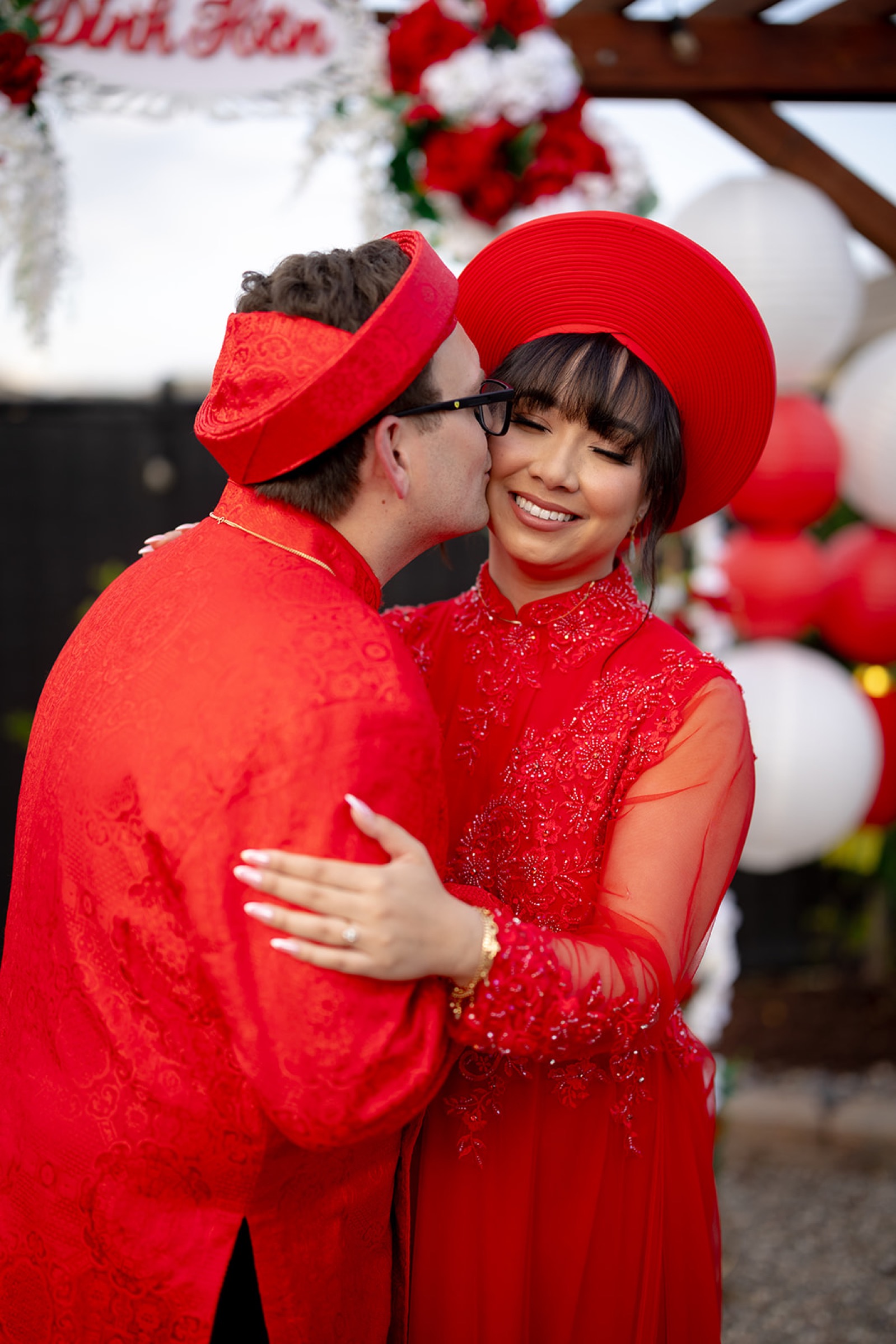 Ryan kissing Janet on the cheek during the tea ceremony, both wearing vibrant red ao dai